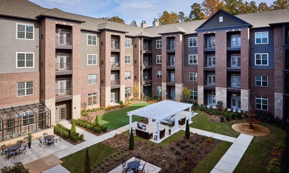 Outdoor courtyard area of a senior living facility with a white pergola covering seating arrangements, surrounded by a well-maintained garden, pathways, and a multi-story brick and siding building in the background.