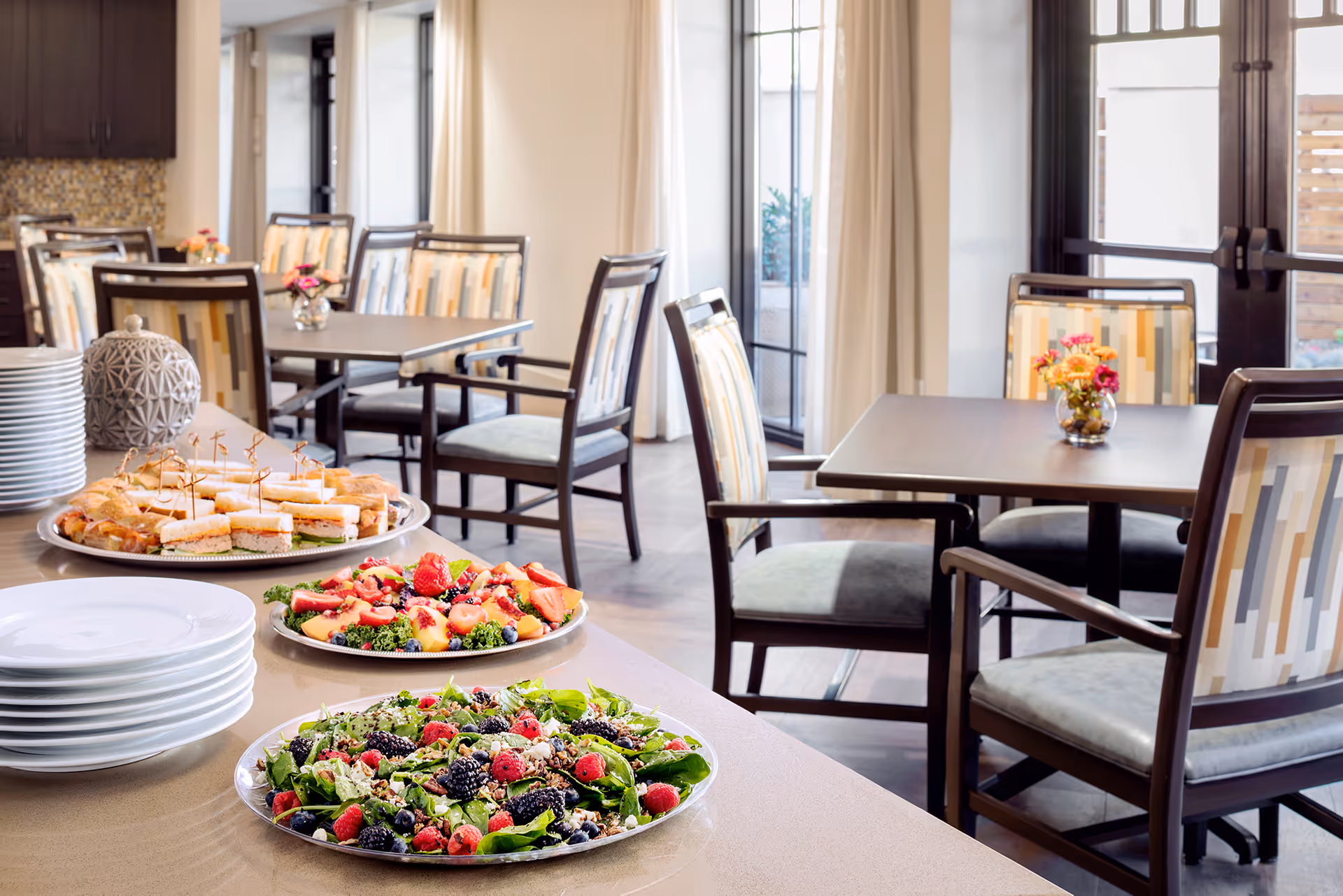 A dining area with several tables and chairs featuring patterned upholstery. On a counter in the foreground, there are plates of fresh salads with berries and a platter of sandwiches. The room has large windows with curtains and a bright, inviting atmosphere.