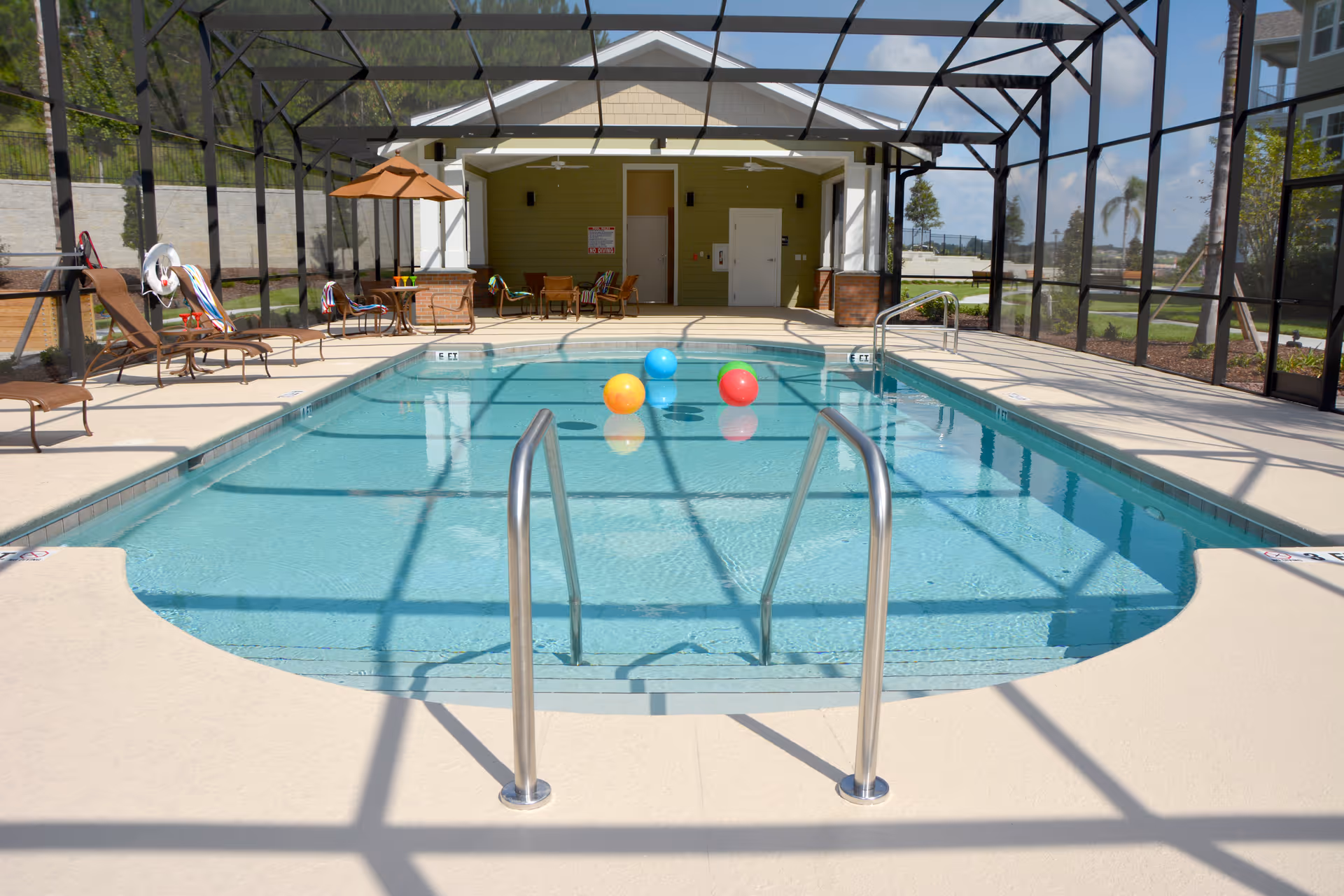 Indoor swimming pool with clear blue water and colorful beach balls floating on the surface. The pool area is enclosed with a screened structure and has lounge chairs, tables with umbrellas, and a small building with doors in the background.