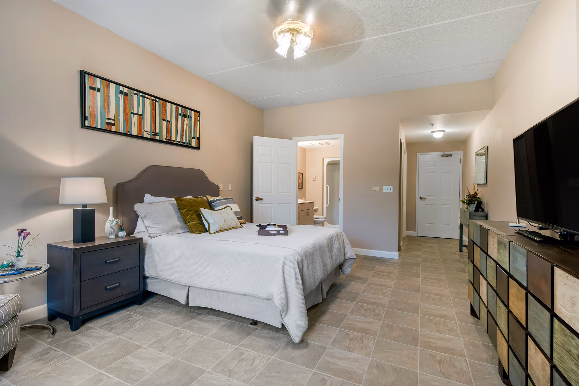 A neatly arranged bedroom in a senior living facility with a bed featuring white bedding and multiple pillows. There is a nightstand with a lamp and decorative items on the left side of the bed. A colorful abstract painting hangs above the bed. The room has tiled flooring and beige walls. A dresser with a large flat-screen TV is on the right side. An open door reveals a bathroom with a shower, and a hallway leads to a closed white door at the back.