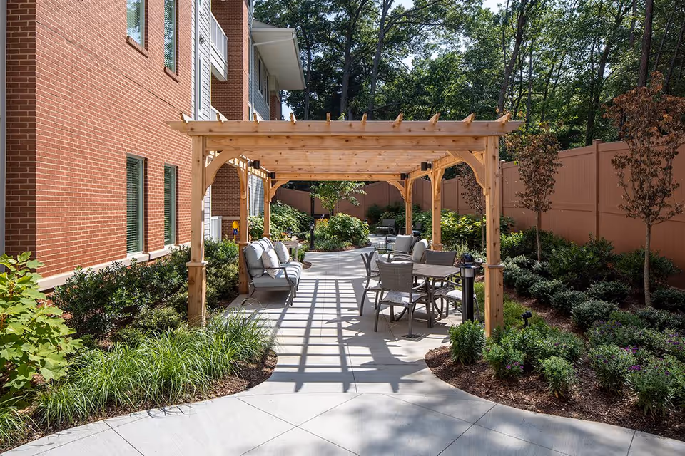 Outdoor seating area with a wooden pergola providing partial shade over cushioned chairs and a table, surrounded by landscaped greenery and a brick building on the left side.