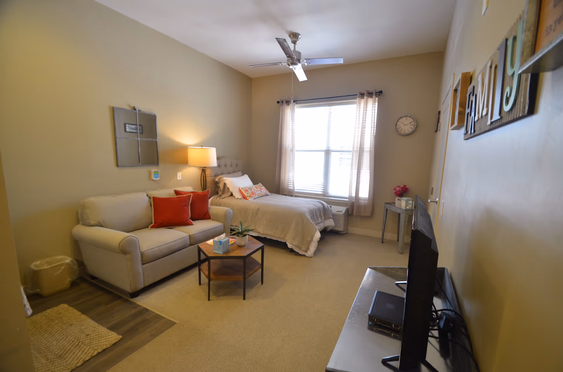 A cozy senior living bedroom featuring a single bed with beige bedding and a colorful pillow, a beige loveseat with two red throw pillows, a small wooden coffee table with a tissue box and plant, a floor lamp, a wall clock, and a TV on a stand. The room has beige walls, a ceiling fan, and a window with sheer curtains letting in natural light.