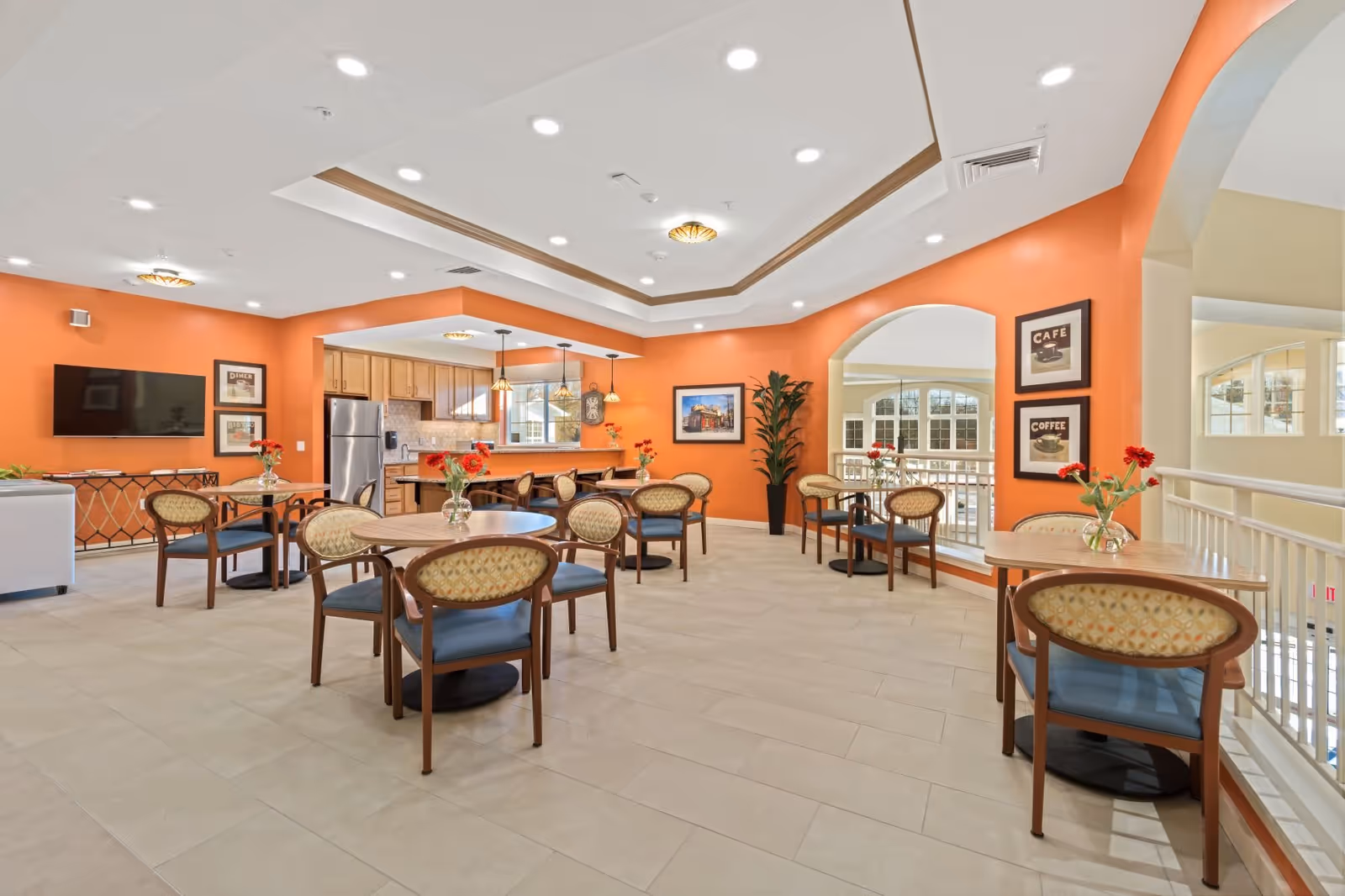 A bright and spacious common dining area with several round tables and chairs, each table decorated with a small vase of red flowers. The walls are painted orange, and there is a kitchen area with wooden cabinets and stainless steel appliances in the background. The room features recessed lighting, framed pictures on the walls, and large windows allowing natural light to enter.