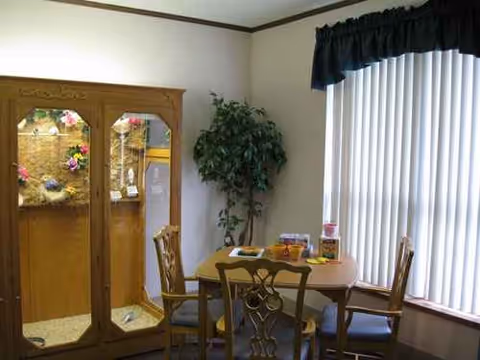 Small dining area in a senior living facility with a cabinet, plant, and blinds.