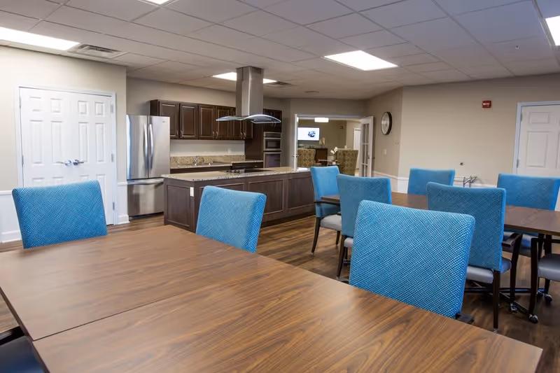 Interior view of a dining area with wooden tables and blue upholstered chairs. In the background, there is a kitchen with dark wood cabinets, a stainless steel refrigerator, and a kitchen island with a range hood above it. The room has beige walls and a ceiling with recessed lighting.