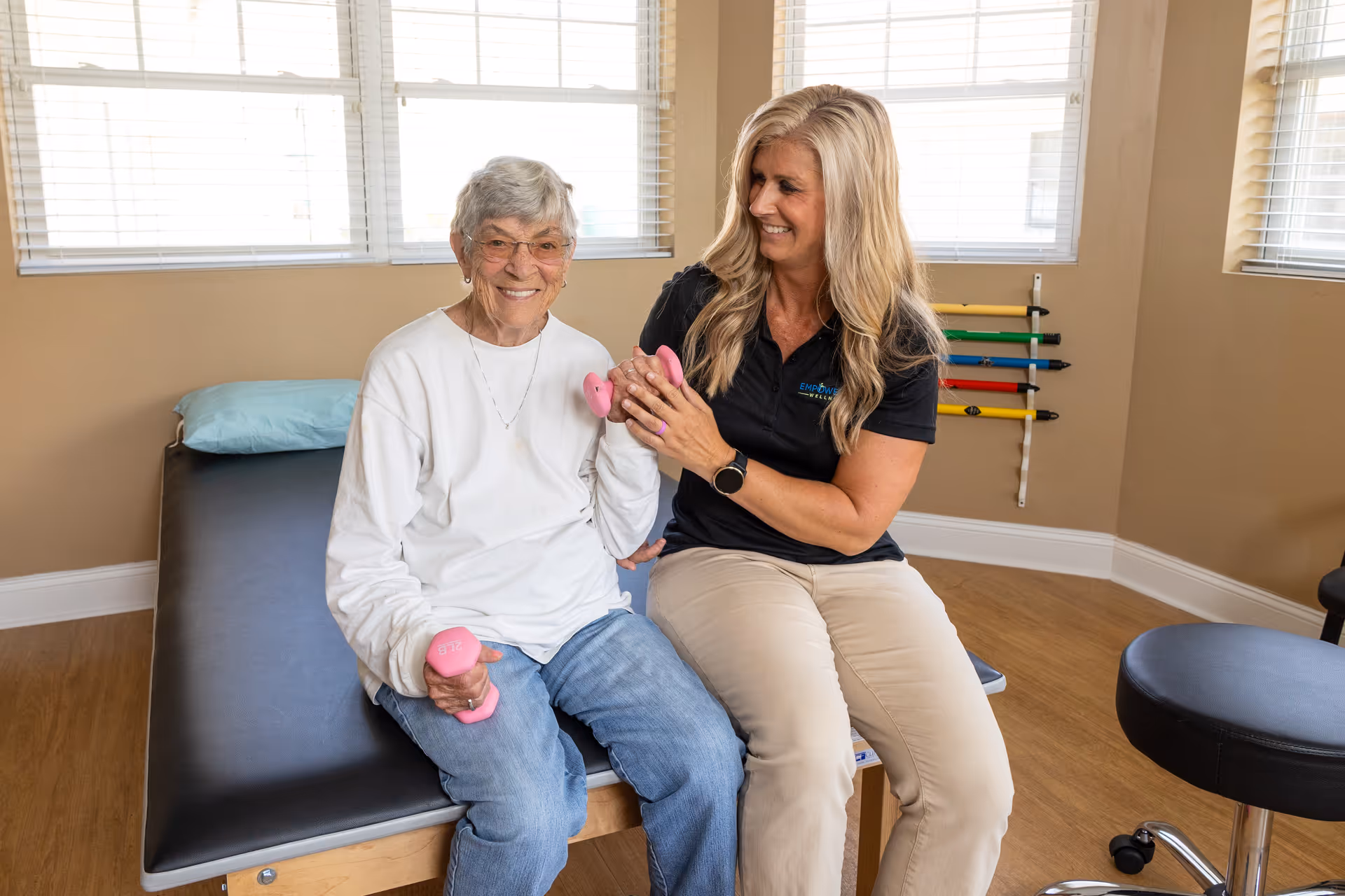 An elderly woman sitting on a padded examination table holding pink dumbbells, smiling, while a female caregiver or therapist in a black polo shirt and beige pants supports her arm in a bright room with windows and exercise equipment on the wall.