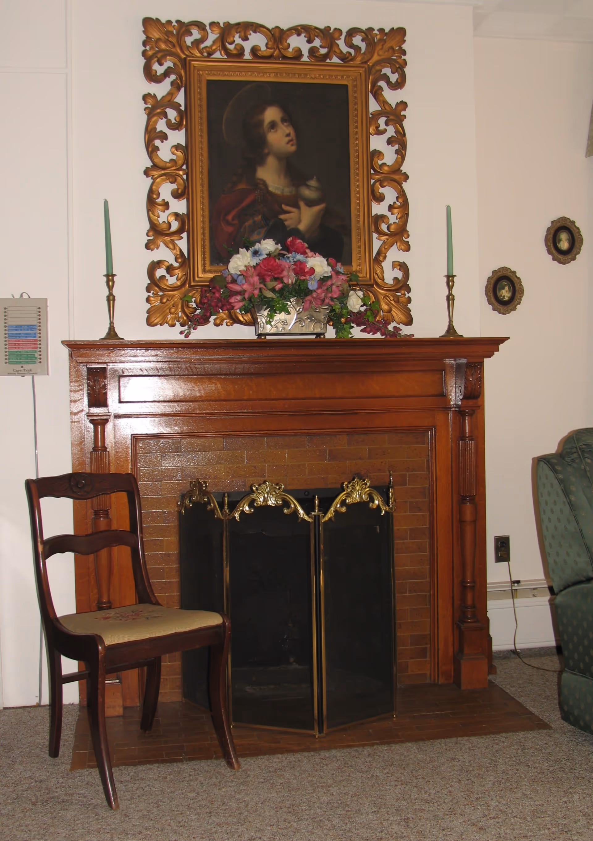 A cozy living room corner featuring a wooden fireplace with a decorative brass screen. On the mantel, there is a floral arrangement and two tall green candles in brass holders. Above the mantel hangs a framed painting of a woman looking upwards, surrounded by an ornate gold frame. To the left of the fireplace is a wooden chair with a cushioned seat, and to the right, part of a green upholstered armchair is visible. The walls are light-colored with small framed pictures on the right side.