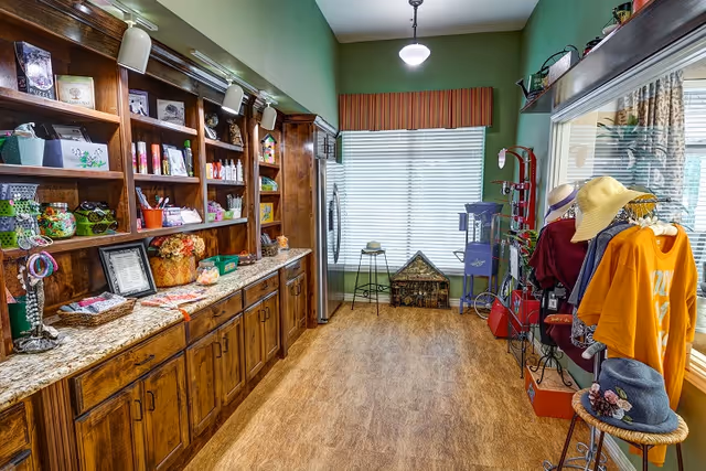 Interior view of a small retail or gift shop area within a facility, featuring wooden shelves stocked with various items such as snacks, accessories, and decorative pieces. There is a granite countertop below the shelves, a stainless steel refrigerator, a popcorn machine, and a display rack with hats and clothing near a large window with blinds. The walls are painted green and the floor has a wood-like finish.