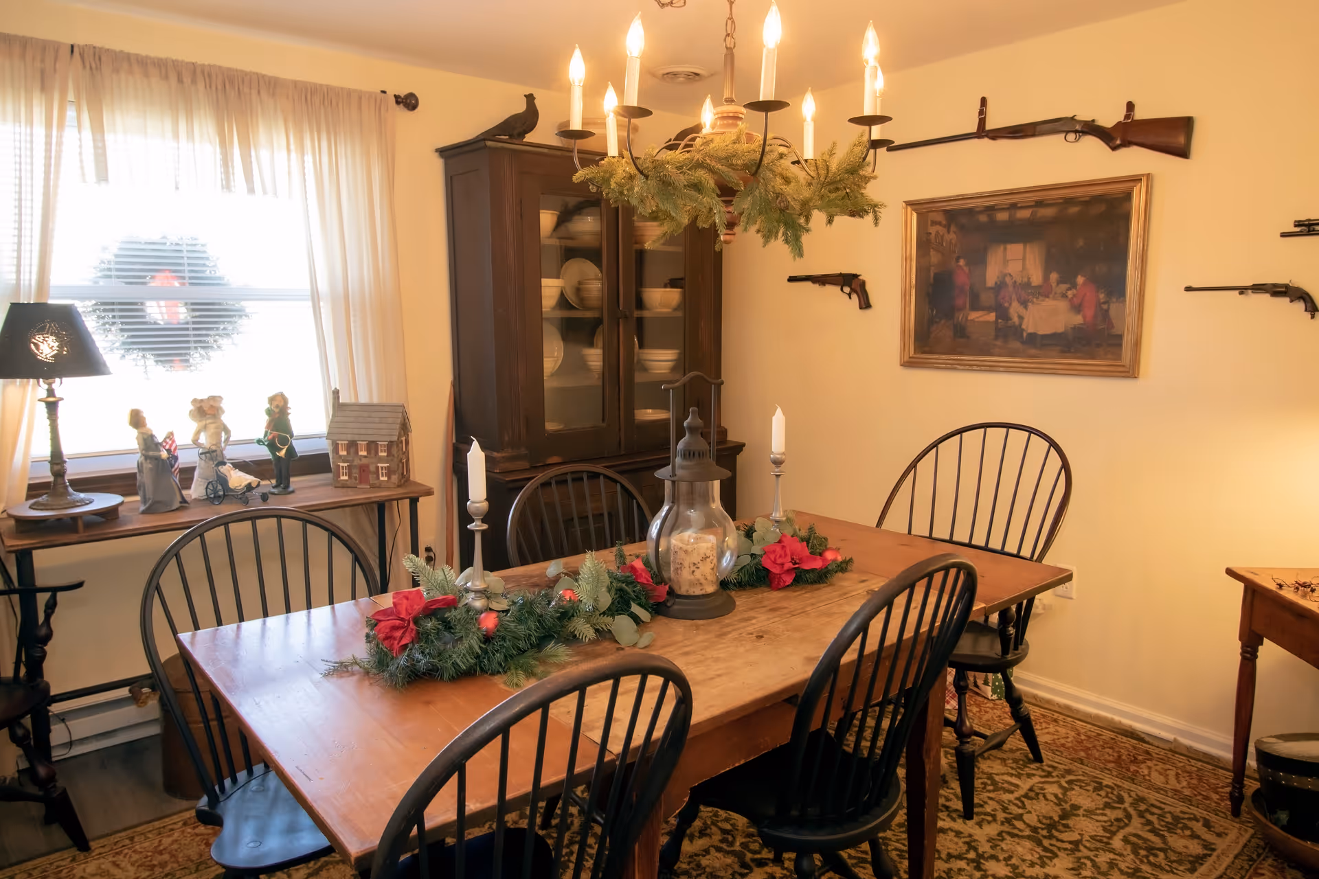A cozy dining room with a wooden table surrounded by six black chairs. The table is decorated with a festive garland, candles, and a lantern. A wooden cabinet with dishes is against the wall, and a window with sheer curtains lets in natural light. The walls feature a framed painting and antique rifles mounted as decoration. A small table near the window holds figurines and a lamp.