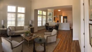 A bright and spacious living room area with large windows featuring white shutters. The room contains a beige sofa, two beige armchairs, a round wooden coffee table with decorative items, and a dining table with chairs in the background. The floor is wooden, and the walls are painted light beige.