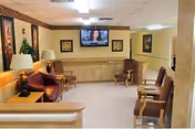A nursing home common area with several chairs arranged along the walls and around small tables. There are two table lamps on side tables, framed artwork on the walls, and a flat-screen TV mounted on the far wall showing a news program. The room has beige walls, a carpeted floor, and ceiling lights providing illumination.