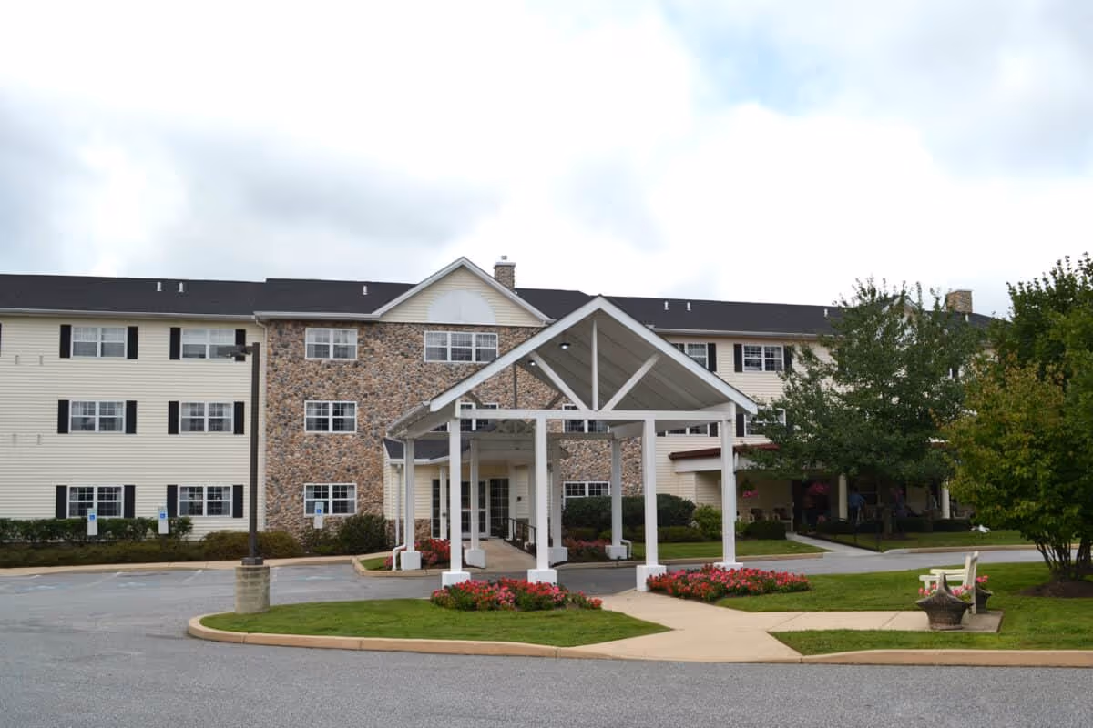 Front exterior of a three-story senior living building showing a covered porte-cochere entrance, landscaped flowerbeds, and a driveway.