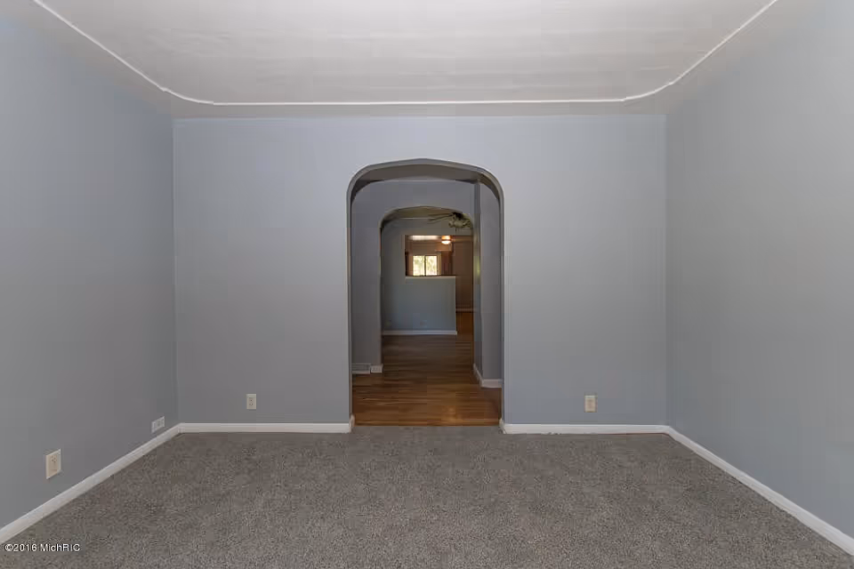 Empty carpeted living room with light gray walls and an arched doorway leading to a hardwood-floored room.