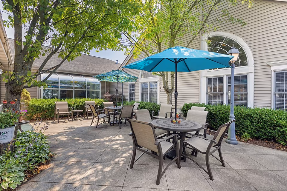 Outdoor patio area at Brookdale Portage Memory Care with round tables and chairs under blue umbrellas, surrounded by greenery and trees next to a beige building with large windows.