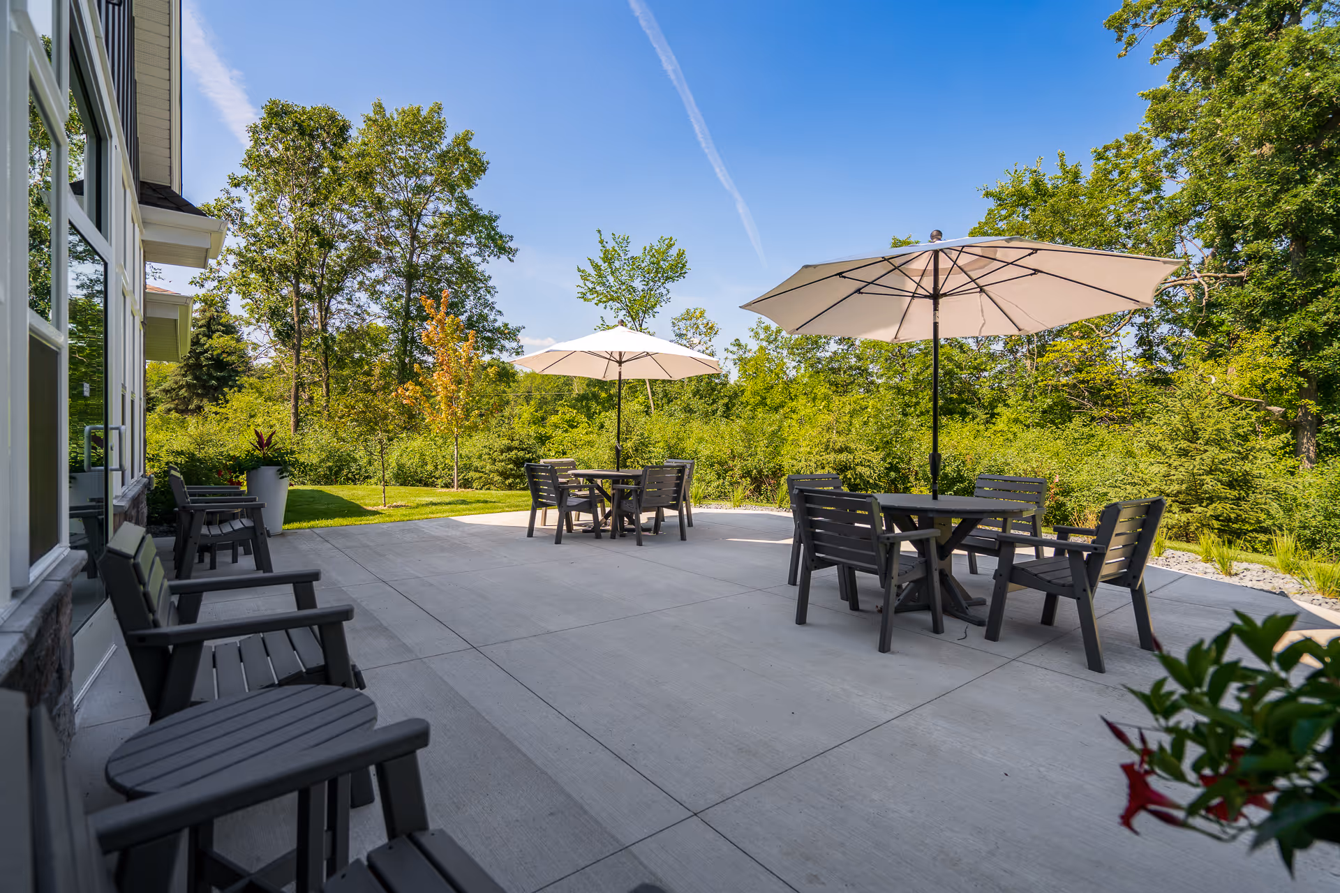 Outdoor patio area at NorBella Senior Living - Champlin with several black chairs and tables under large beige umbrellas, surrounded by green trees and plants under a clear blue sky.