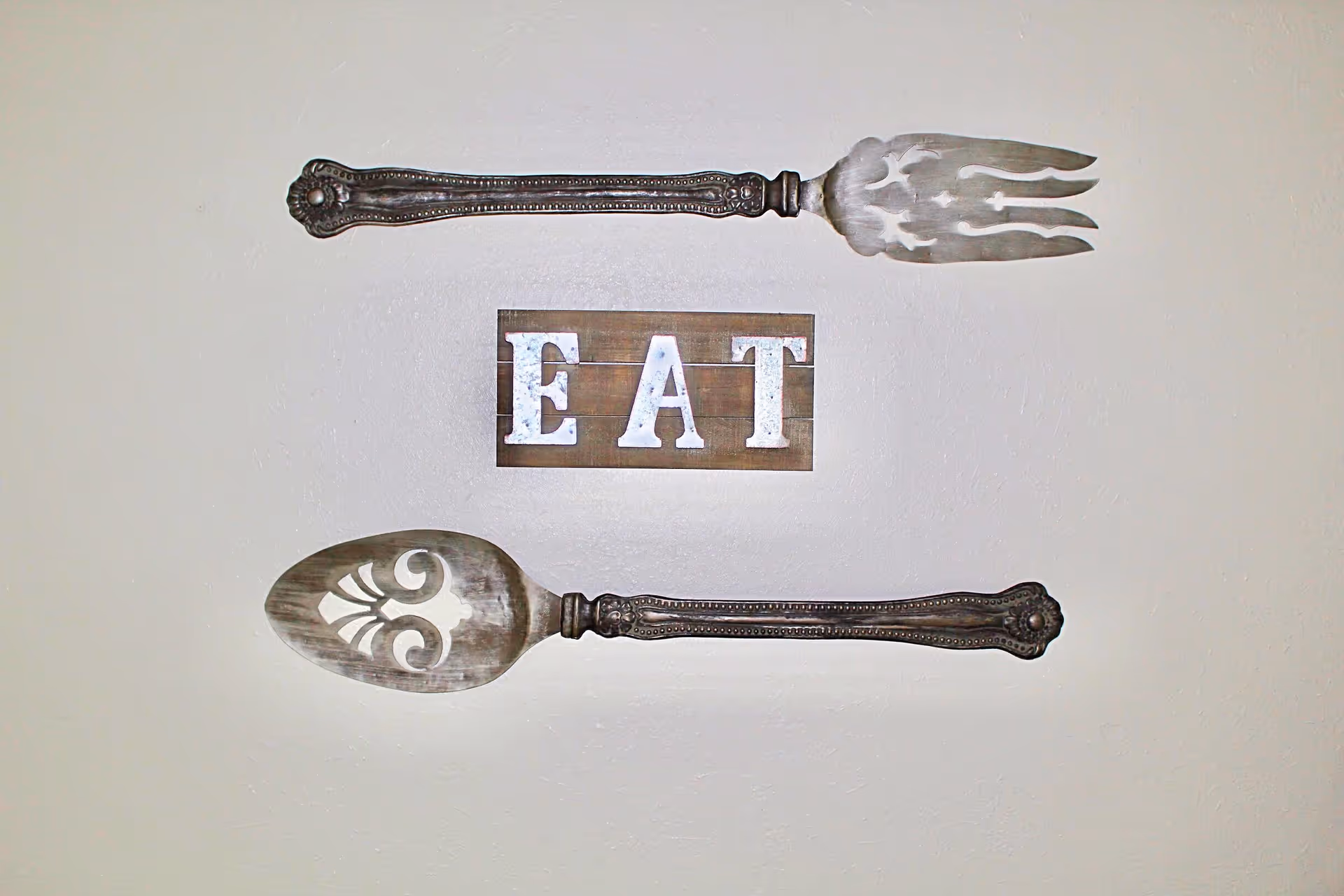 Wall decoration featuring a large vintage-style fork and spoon with intricate designs, positioned horizontally above and below a wooden sign with the word 'EAT' in white letters.