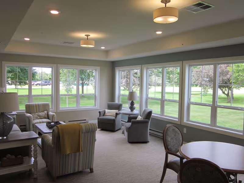 A bright and spacious living room in a senior living facility with large windows showing a green outdoor view. The room contains several comfortable armchairs, a small round table with chairs, two table lamps, and ceiling lights. The walls are painted light gray and the carpet is beige.