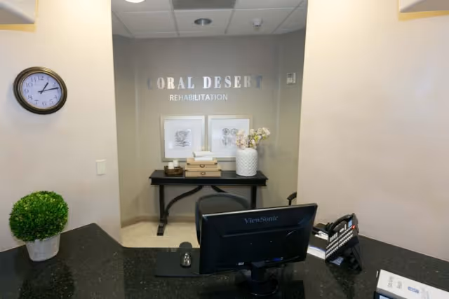 Reception area of Coral Desert Rehabilitation featuring a black desk with a computer monitor, a mouse, a phone, and some papers. Behind the desk is a wall with the facility name 'Coral Desert Rehabilitation' displayed, a black table with decorative boxes, framed artwork, and a white vase with flowers. A round wall clock and a small green potted plant are also visible.