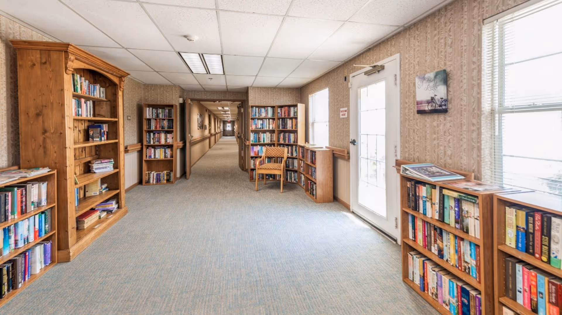 Well-lit interior hallway lined with bookshelves and a reading chair, leading down a long corridor with a door and windows on the right.