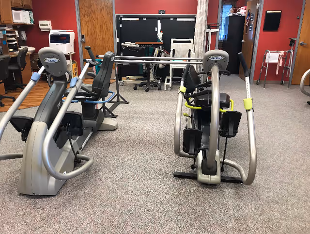 Interior room with exercise equipment including two recumbent exercise bikes and other physical therapy equipment, with office furniture and medical supplies visible in the background.