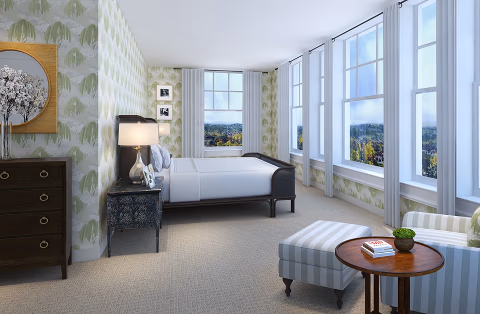 A bright and spacious bedroom with large windows along one wall, allowing natural light to fill the room. The room features a bed with white bedding, a bedside table with a lamp and picture frames, a dark wooden dresser with a round mirror above it, and a seating area with a striped ottoman and a small round wooden table holding a plant and books. The walls have a green and white patterned wallpaper.