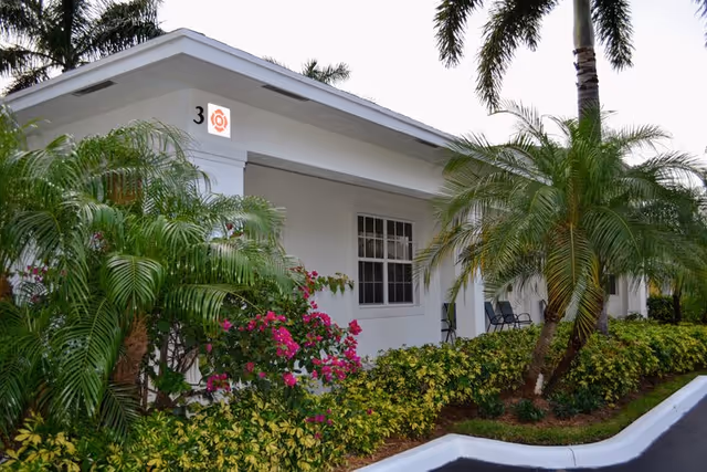 Exterior view of a single-story white building with a window and a small porch area. The building is surrounded by lush green plants, palm trees, and flowering bushes. The number 3 is visible on the corner of the building.