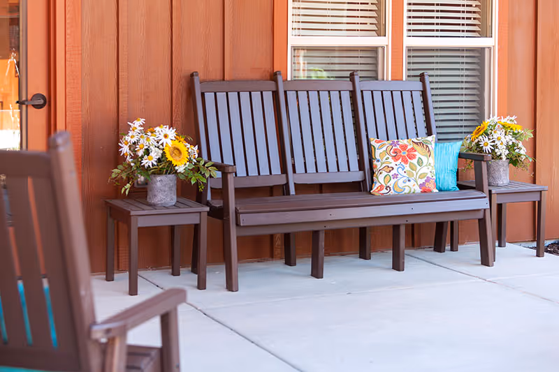 Three-seat wooden bench with colorful pillows and potted flowers on a porch in front of brown siding and windows.