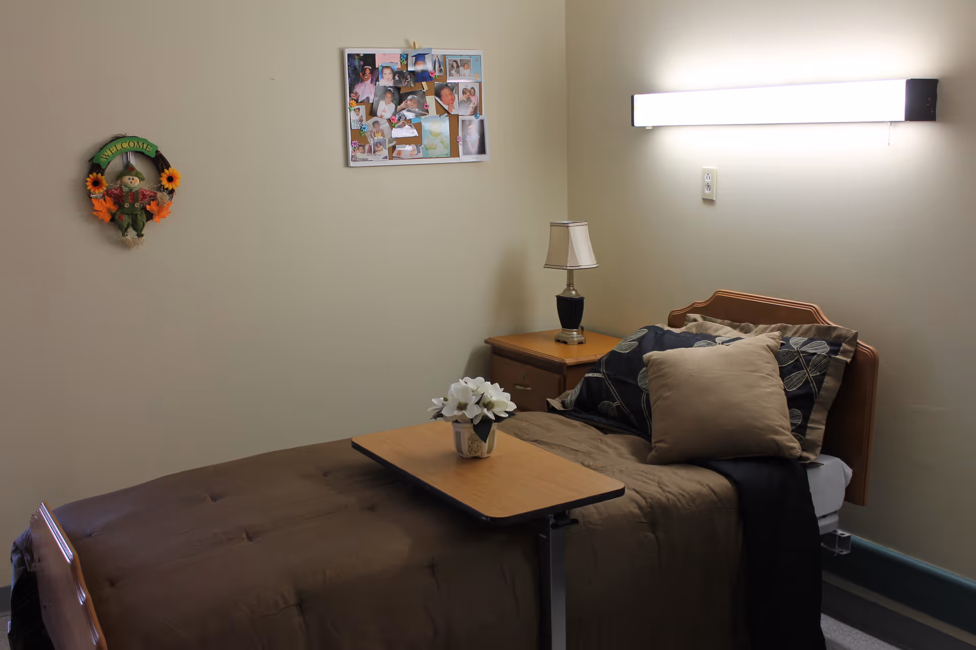 Small furnished bedroom with a single bed, overbed table with flowers, nightstand and lamp, and photos and wall decor.