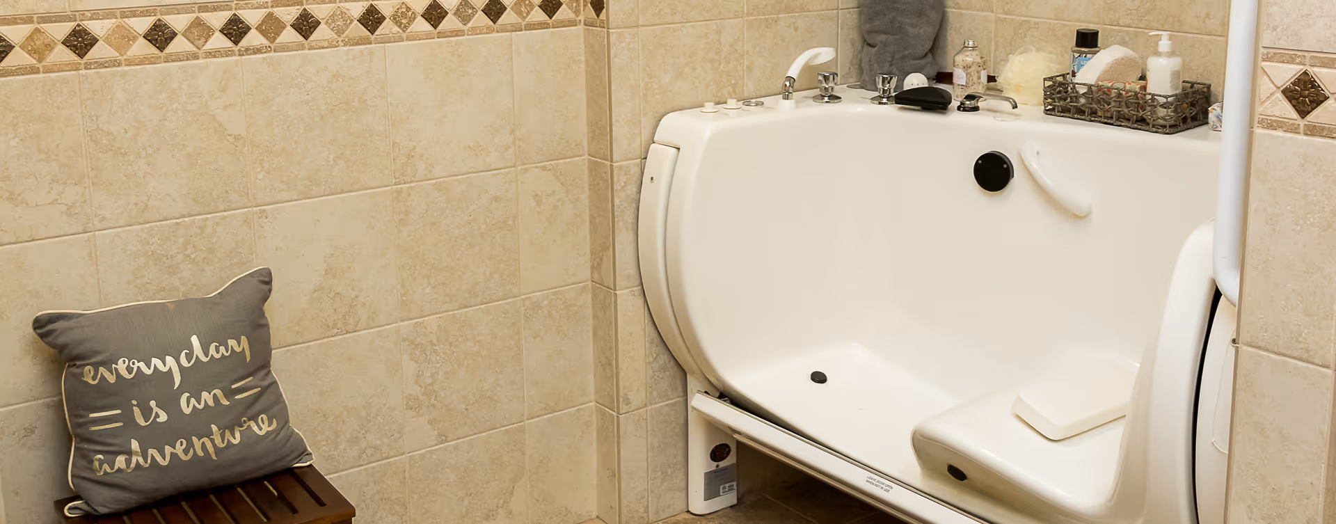 A walk-in bathtub with a built-in seat and safety handles in a tiled bathroom. On the left, there is a small wooden stool with a gray pillow that has the words 'every day is an adventure' written on it. Various bath items are placed on the edge of the tub.
