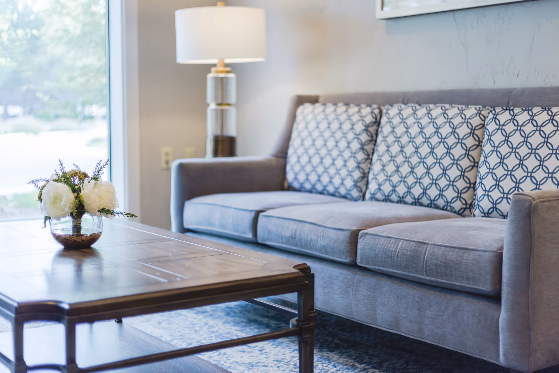 A cozy living room area with a gray sofa adorned with patterned cushions, a wooden coffee table with a small glass vase containing white flowers, a standing lamp with a white shade, and a large window letting in natural light.