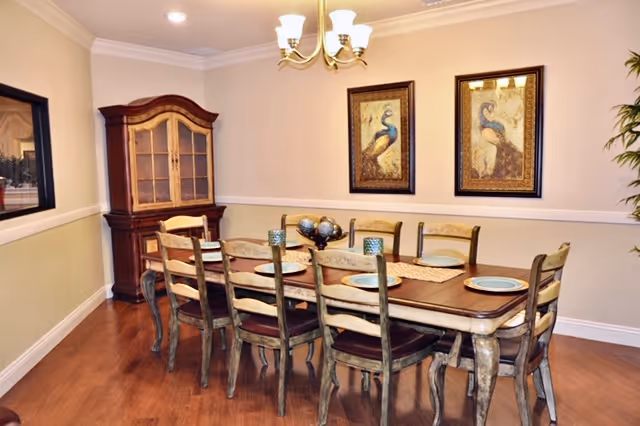 A dining room with a long wooden table set with eight chairs. The table has placemats and decorative items on it. On the wall behind the table are two framed paintings of peacocks. A wooden china cabinet is positioned in the corner, and a chandelier with five lights hangs from the ceiling.
