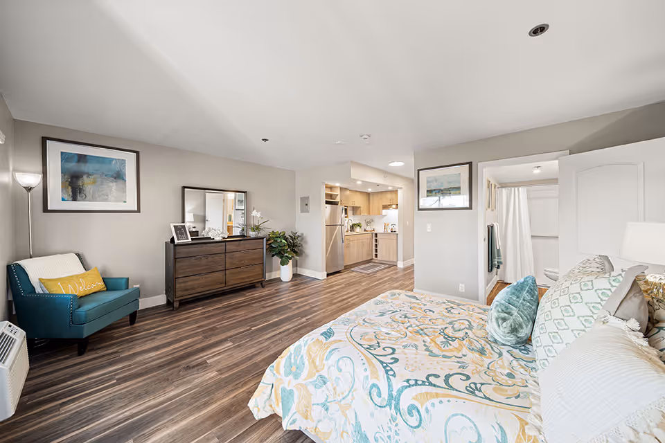 A spacious senior living facility room at Ivy Park of Wellington featuring a bed with patterned bedding, a teal armchair with a yellow pillow, a dark wooden dresser with a mirror, a potted plant, and a kitchenette with stainless steel appliances. The room has wood flooring and light-colored walls, with a bathroom visible through an open door.