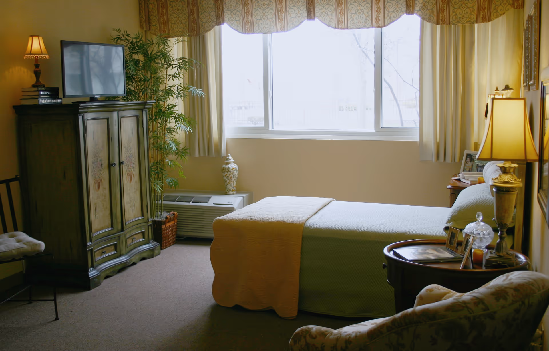 A cozy bedroom in a retirement community featuring a single bed with a yellow and green quilt, a wooden nightstand with a lamp and framed photos, a cushioned armchair, a wooden cabinet with a TV on top, a potted plant, and a large window with curtains letting in natural light.
