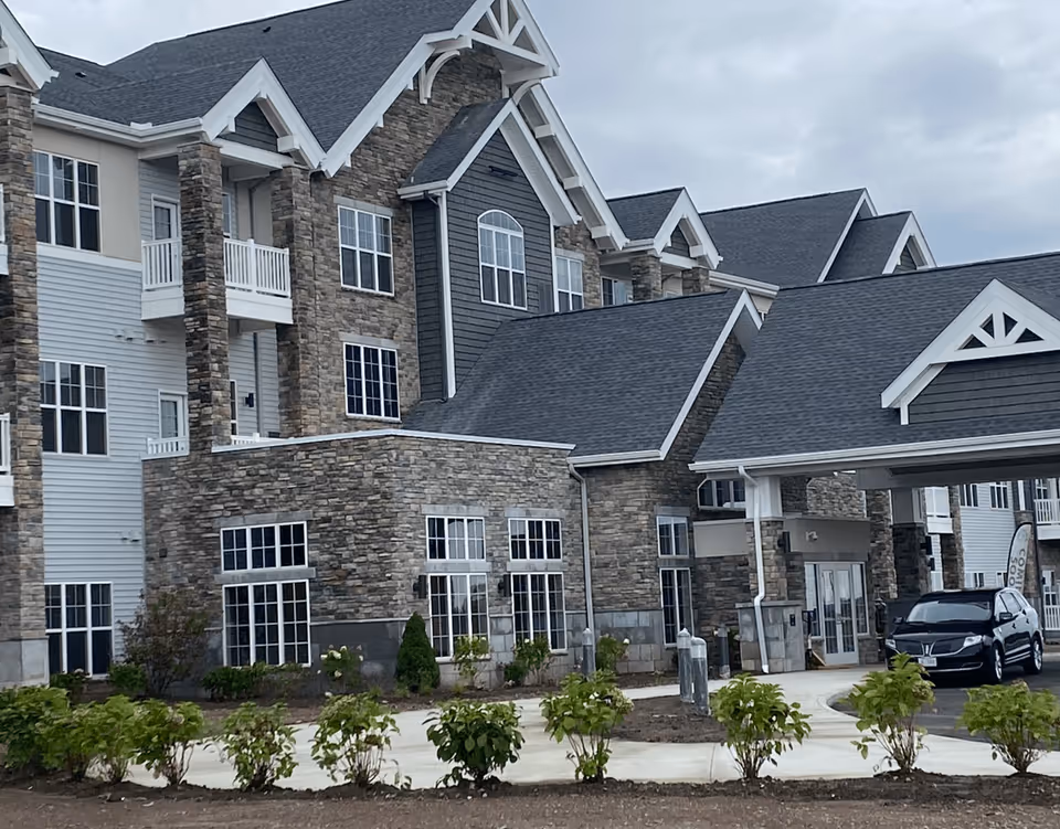 Exterior view of a multi-story senior living facility building with stone and siding facade, multiple windows, balconies, and a covered entrance with a black SUV parked nearby. There are small bushes and a paved walkway in front of the building under a cloudy sky.