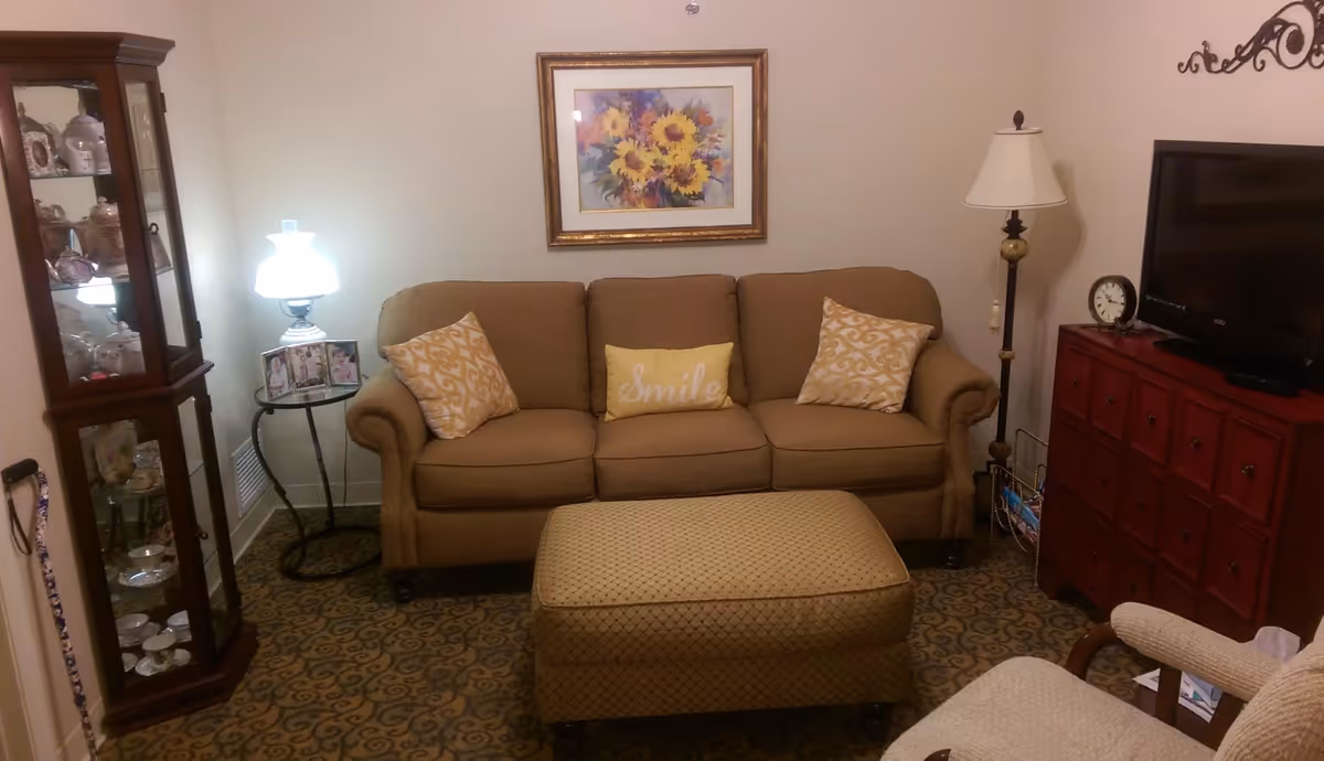Cozy living room featuring a beige sofa with decorative pillows, a matching ottoman, lamps, TV on a red cabinet, and a glass display cabinet.