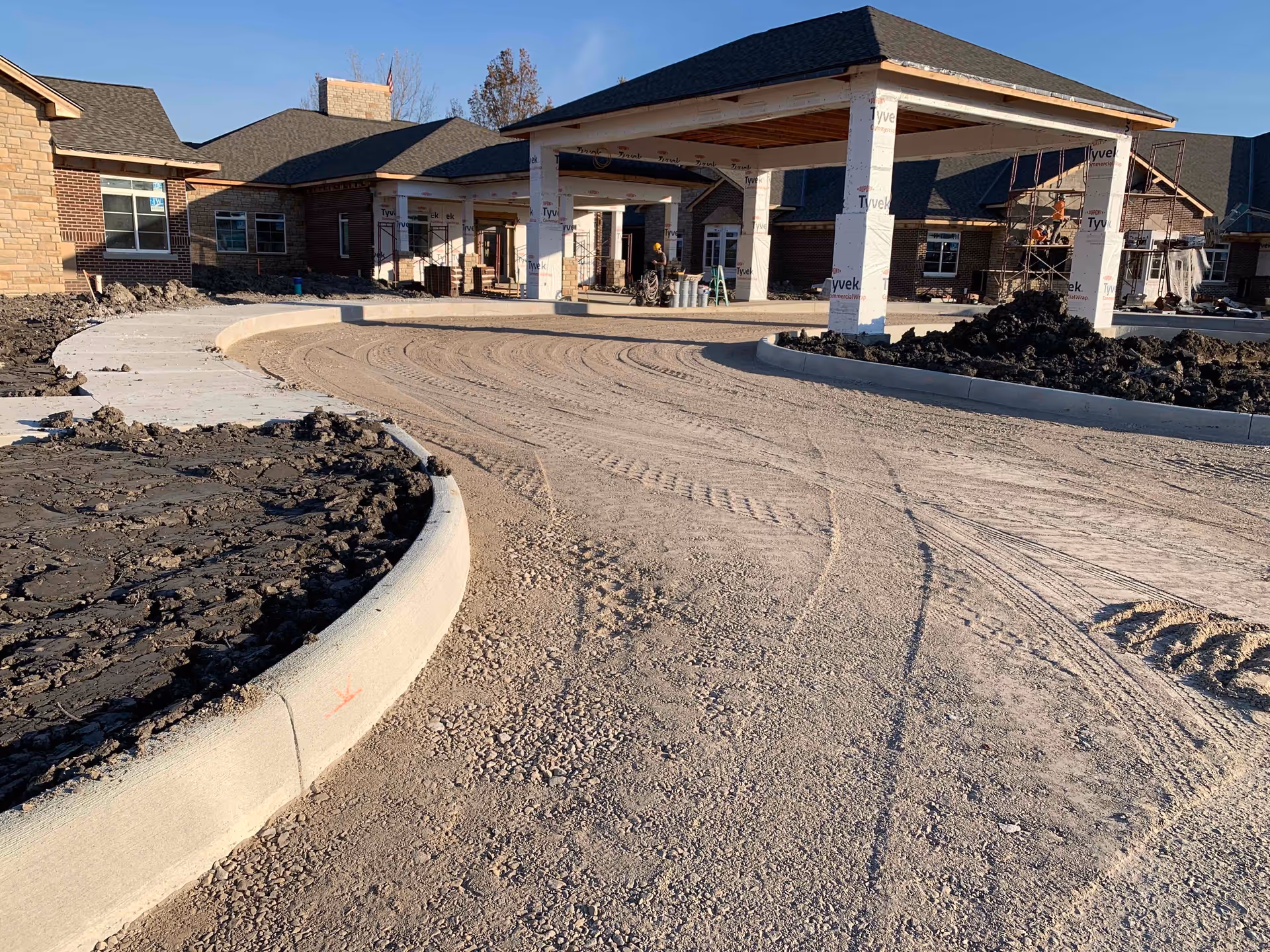 View of the front entrance of The Ganzhorn Suites facility under construction, showing a curved driveway with fresh gravel, concrete curbs, and a covered drop-off area supported by columns wrapped in Tyvek material. The building exterior features brick and stone walls with windows, and construction materials and workers are visible in the background.