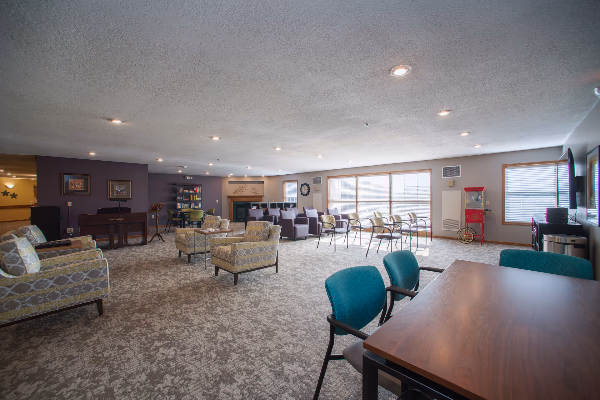 A spacious senior living common area with multiple seating options including patterned armchairs, purple chairs, and green chairs around tables. The room has large windows letting in natural light, a fireplace with decorative artwork above it, a bookshelf, a popcorn machine, and a TV mounted on the wall.