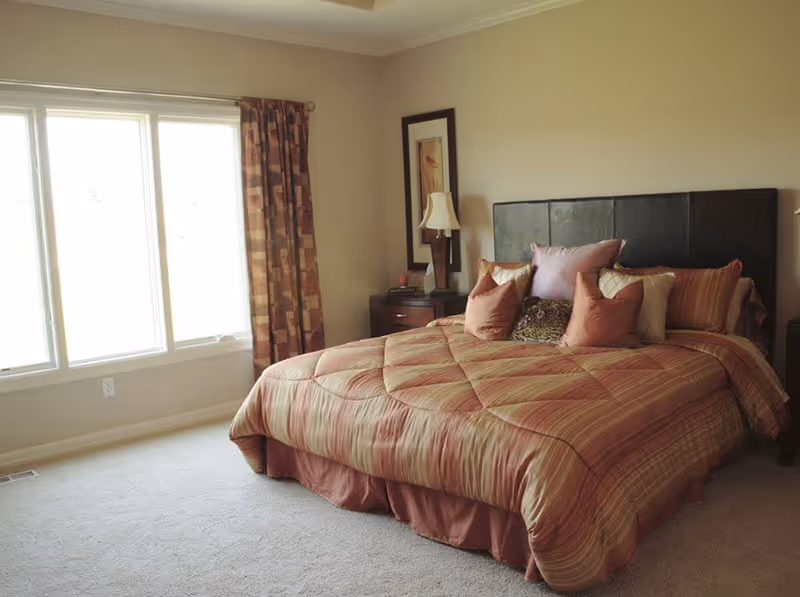 Sunlit bedroom with a large bed dressed in striped bedding, nightstands with a lamp, and a window with patterned curtains.