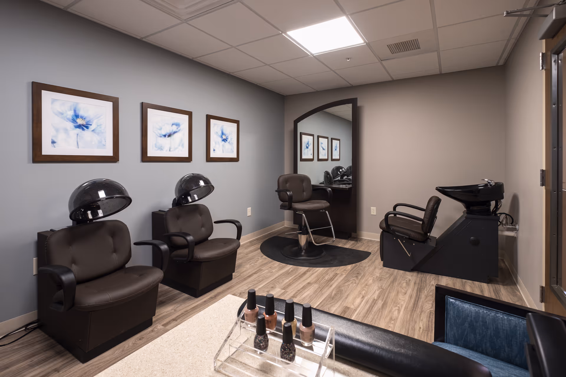 Interior view of a salon area in a senior living facility featuring two black hair drying chairs with hoods, a styling chair in front of a large mirror, and a black hair washing station. The walls are painted gray with three framed floral artworks. A table with nail polish bottles is visible in the foreground.