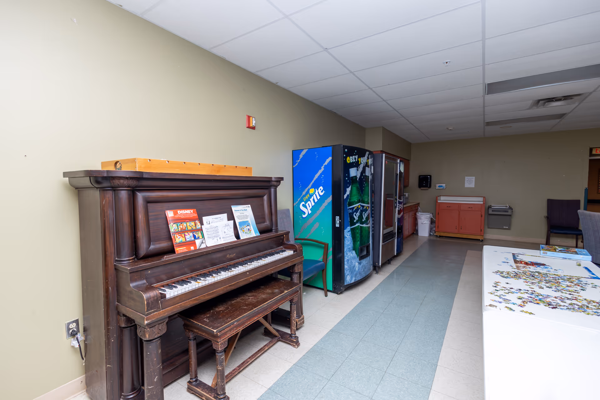 A common room with an upright piano, vending machines, a puzzle table and seating.