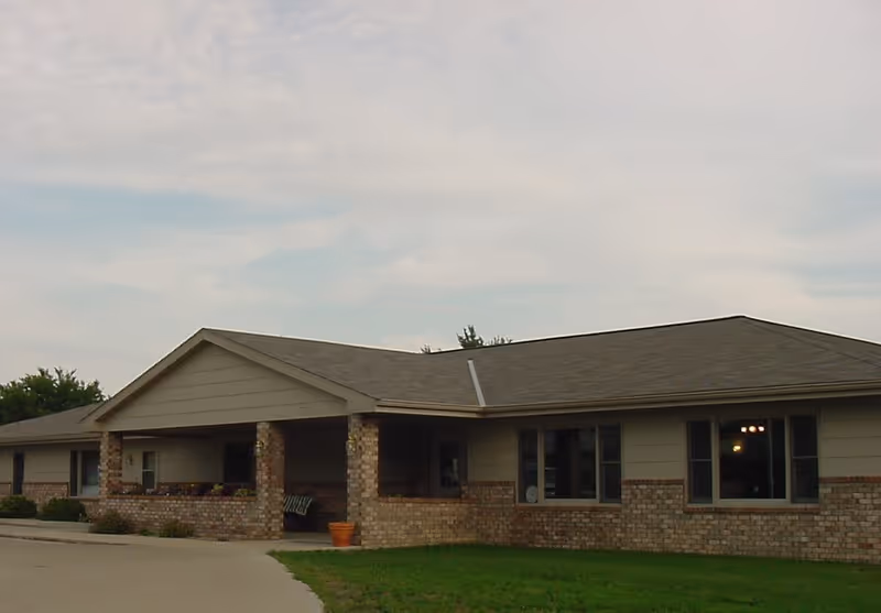 Single-story building with a brick and siding exterior, a covered porch area with pillars, and several windows. The building is surrounded by a driveway and a small grassy area under a partly cloudy sky.