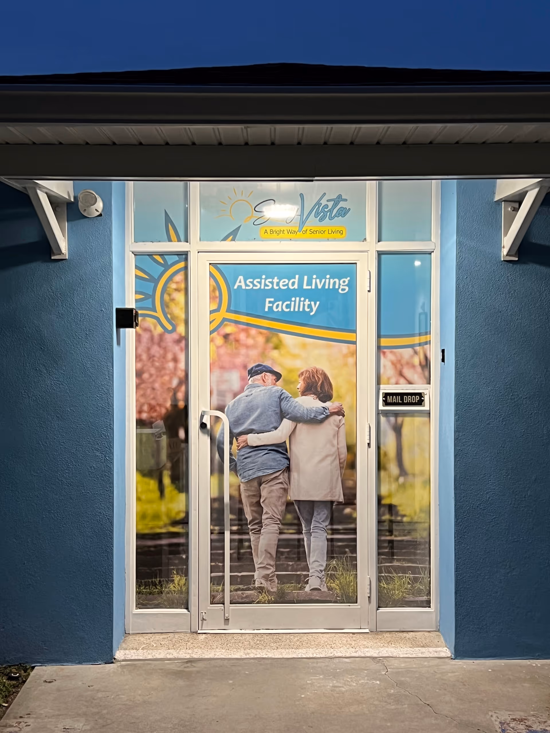 Entrance door of Sunny Vista Assisted Living Facility LLC with a large graphic showing an elderly couple walking arm in arm outdoors. The door has text that reads 'Assisted Living Facility' and a mail drop slot on the right side. The building exterior is painted blue.