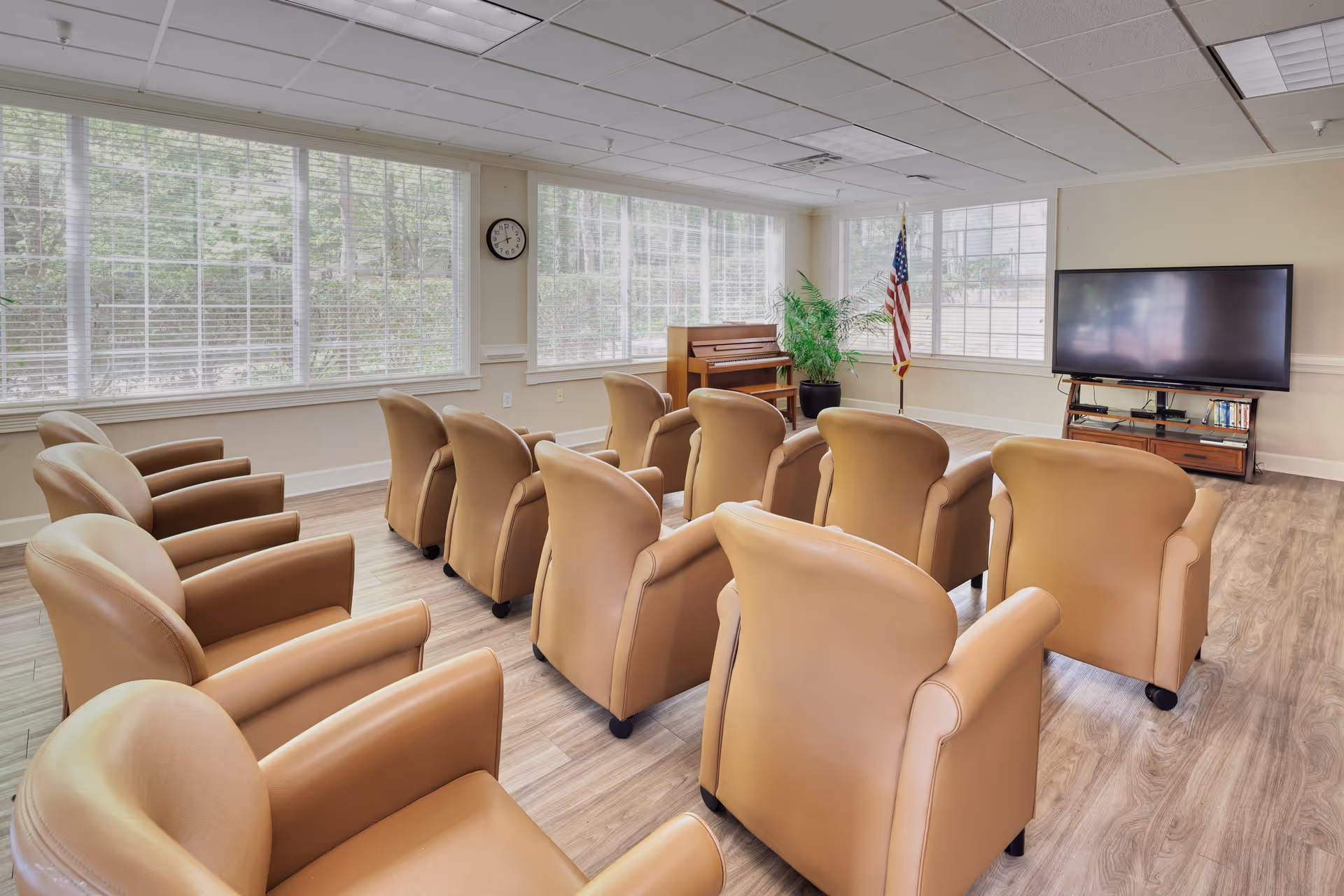 A bright room with large windows covered by white blinds, featuring three rows of tan leather armchairs facing a flat-screen TV on a wooden stand. There is a piano and an American flag in the corner, with a potted plant nearby. The floor is wood, and a clock is mounted on the wall between the windows.
