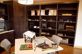 A cozy interior room with a white table in the center surrounded by four chairs. On the table are various items including a book on a stand, a small tray with compartments, and some rolled-up scrolls. Behind the table is a large dark wooden shelving unit filled with decorative items, books, and rolled papers. The room has warm lighting and wooden flooring.