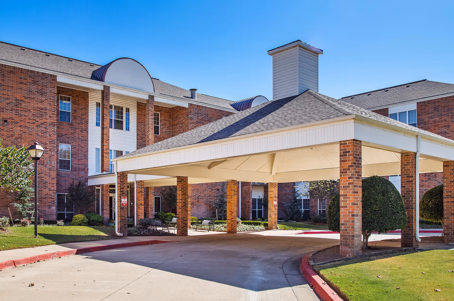Exterior view of a brick senior living facility with a covered driveway entrance supported by brick pillars, manicured bushes, and a clear blue sky.