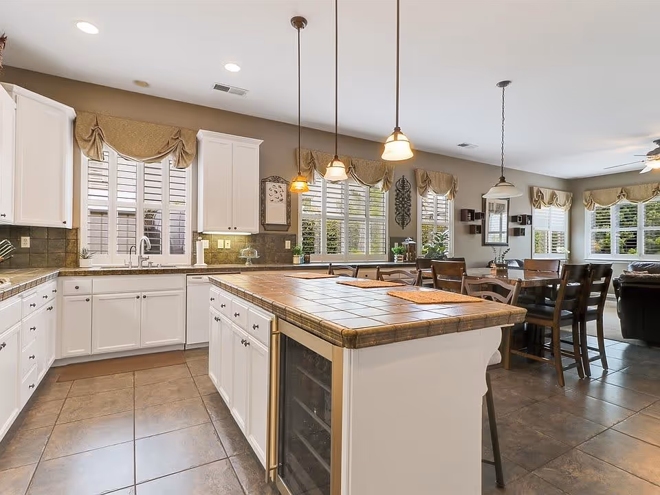 Bright and spacious kitchen with white cabinets, a large tiled island with bar stools, pendant lighting, and multiple windows with beige valances. The kitchen opens into a dining area with a wooden dining table and chairs, and a living room with a ceiling fan and large windows.