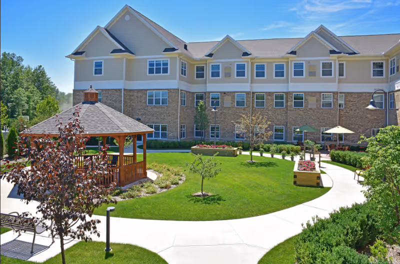 Outdoor courtyard area of a senior living facility with a wooden gazebo, green lawn, small trees, flower beds, benches, and a paved walking path. The three-story building with beige siding and brick exterior surrounds the courtyard under a clear blue sky.