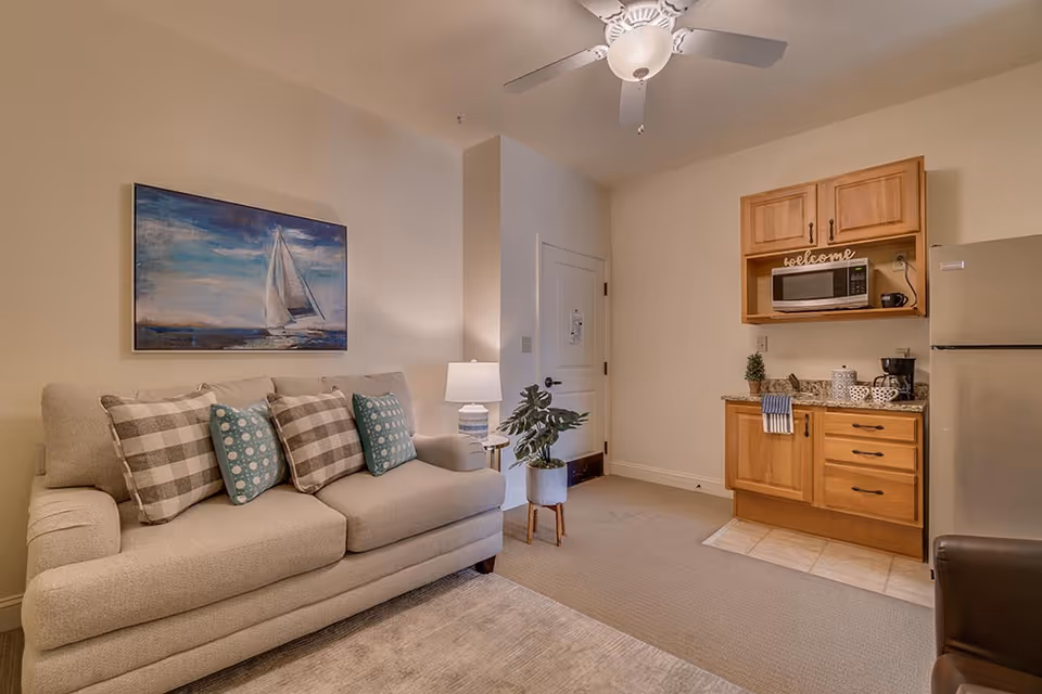 A cozy living area with a beige sofa adorned with patterned cushions, a small side table with a lamp, a potted plant, and a kitchenette featuring wooden cabinets, a microwave, a coffee maker, and a refrigerator. A ceiling fan with a light fixture is mounted above, and a painting of a sailboat hangs on the wall above the sofa.
