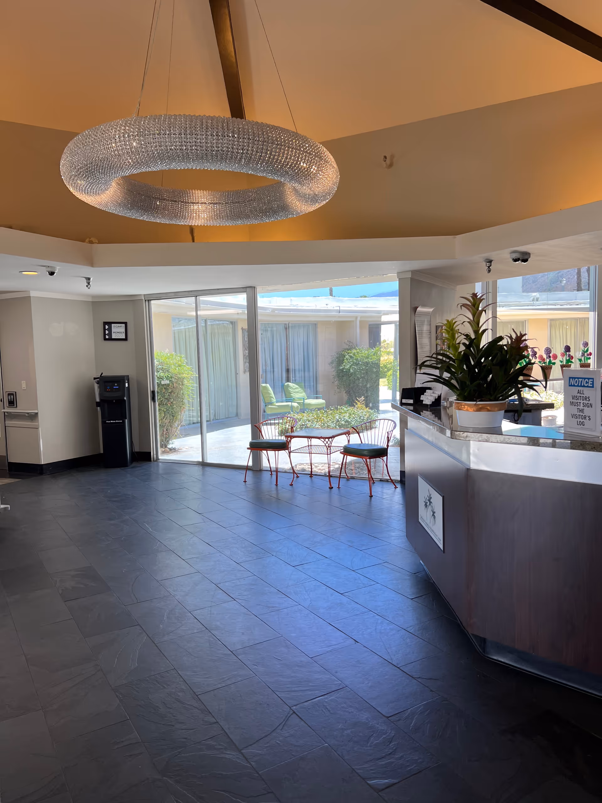 Interior view of a nursing facility lobby with a modern circular chandelier hanging from the ceiling. There is a reception desk with a potted plant and a notice sign. Large glass sliding doors lead to an outdoor courtyard with green chairs and plants. The floor is covered with dark tiles.