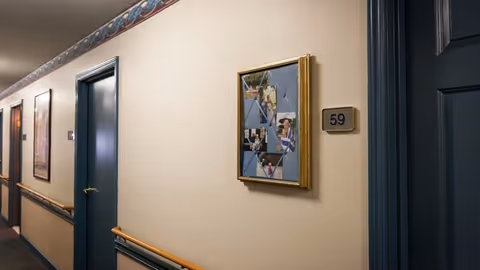 Interior hallway of a senior living facility with beige walls, blue doors, wooden handrails, and framed pictures on the walls. One door is labeled with the number 59.
